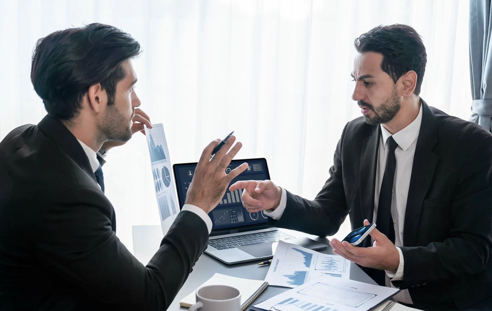 Two businessmen in dark suits sit across from each other at a desk